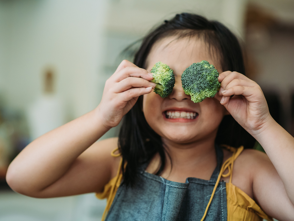smiling little girl holding broccoli in front of her eyes