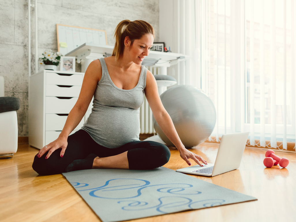 pregnant woman sitting on yoga mat looking at laptop