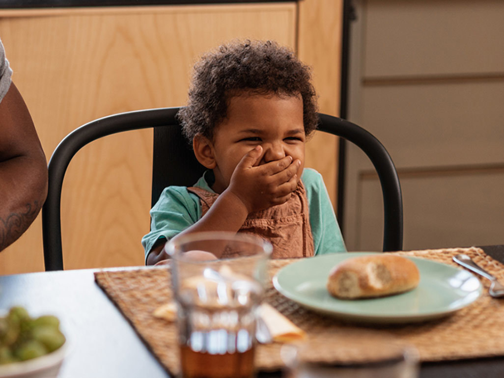 A toddler covering his mouth at a dining table